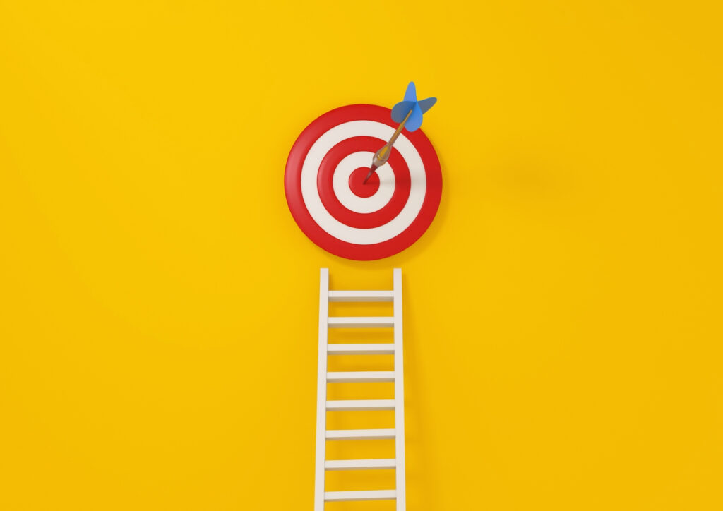 The photograph shows a dartboard on a yellow wall. A ladder is leaning against this dartboard.